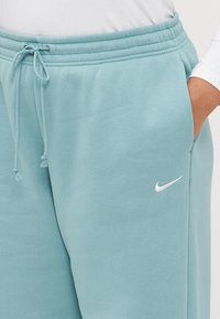 Lyseblå Nike joggebukser med elastisk linning, snor, lommer på siden, og en liten hvit logo foran. Glatt tekstur.