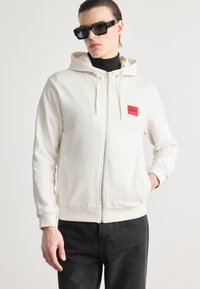 Felpa bianca con zip realizzata in tessuto morbido, caratterizzata da una piccola etichetta rossa sul lato sinistro del petto. Indossata con occhiali da sole neri e jeans scuri.