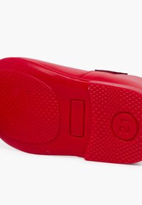 Suela de zapato de goma roja con patrón de agarre texturizado, incluye un botón rectangular y un logo circular con "21". Bordes suaves y forma redondeada.
