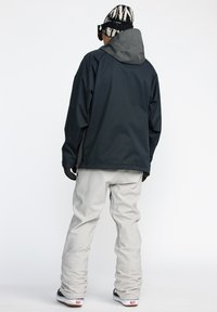 Veste imperméable noire avec capuche grise, associée à un pantalon de neige gris clair. Les caractéristiques incluent une coupe décontractée et un tissu texturé. Bottes de neige visibles.