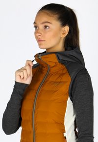 Gilet isolato con corpo arancione, cappuccio nero e maniche grigie. Presenta una chiusura frontale con zip, texture trapuntata e logo sul petto.