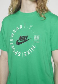 Vihreä puuvillapaita, jossa on keskellä musta Nike-logo, ympyrämainen valkoinen teksti "SPORTSWEAR" ja "SINCE 1972" eri kokoisina sen ympärillä.