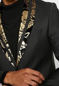 Schwarze Blazer mit gold- und schwarz gemustertem Samt-Revers, strukturiertem Stoff und einem dezenten Taschendetail. Über einem schwarzen Rollkragenpullover getragen.