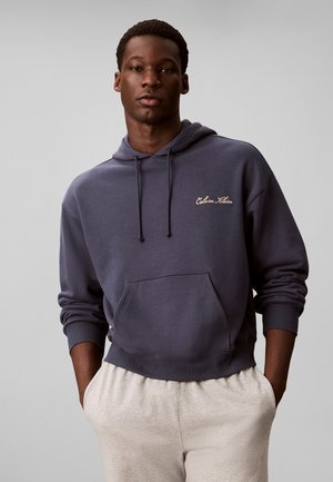 Junger Mann mit dunkelblauem Calvin Klein Hoodie und hellbeigen Hosen, die Hände in den Taschen, stehend vor einem schlichten grauen Hintergrund.