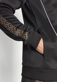 Zwarte jas met een geometrisch gouden accent langs de mouw, met ribgebreide manchetten en een ritszak. Gladde stoffen textuur.