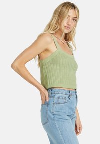 Top corto lavorato a maglia verde con bretelle larghe, caratterizzato da un motivo testurizzato. Indossato con jeans a vita alta di colore azzurro chiaro. Design equilibrato e aderente.
