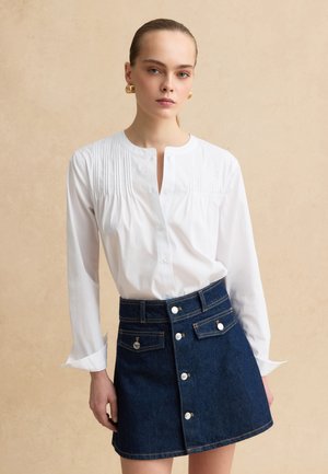 Mujer vistiendo una blusa blanca de manga larga con pliegues y una minifalda de mezclilla azul con botones frontales y bolsillos, posando frente a un fondo beige.