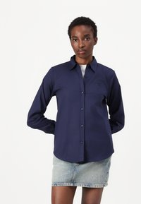 Camicia button-up blu navy con colletto, maniche lunghe e un logo discreto. Abbinata a una mini gonna in denim chiaro, che mette in risalto una texture liscia.