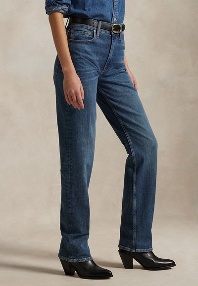 Moteris dėvi tiesias mėlynas džinsines kelnes, juodą diržą, juodus kulniukais aukštakulnius aulinukus ir kišenėje įkištą džinsinę marškinių suvertomis rankovėmis.