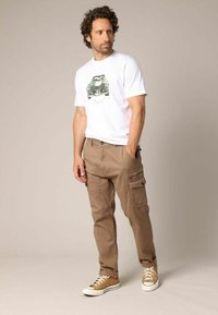 Witte graphic t-shirt met autodesign, bruine cargobroek met meerdere zakken en beige sneakers. Casual outfit met een ontspannen pasvorm.