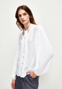 Weiße Bluse mit gefältelten langen Ärmeln, einer Halsband und goldenen Knöpfen vorne. Weicher Stoff mit glatter Textur.