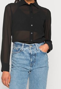 Chemise noire transparente à manches longues avec un col et une fermeture à boutons sur le devant, associée à un jean en denim bleu taille haute. Les boutons sont gris.