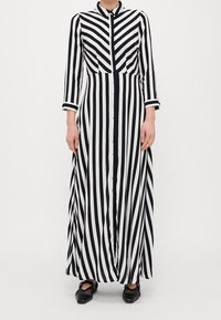 Kobieta ubrana w długą sukienkę maxi w czarno-białe pionowe paski z długim rękawem, zapinaną na guziki z przodu oraz w czarne szpilki.
