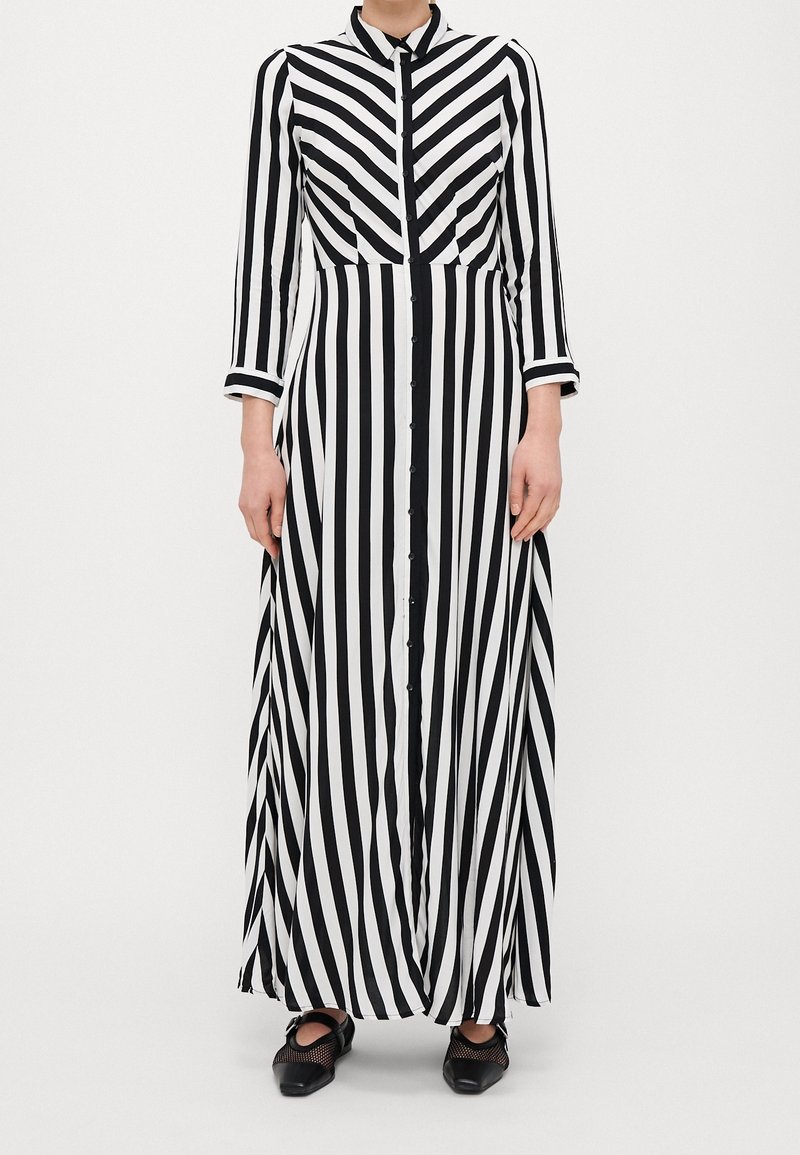 Kobieta ubrana w długą sukienkę maxi w czarno-białe pionowe paski z długim rękawem, zapinaną na guziki z przodu oraz w czarne szpilki.