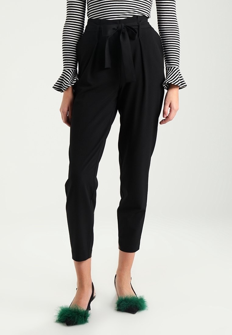 Femme portant un pantalon noir taille haute fuselé avec ceinture à nœud, un haut à manches longues rayé, et des chaussures noires avec des touches de plumes vertes.