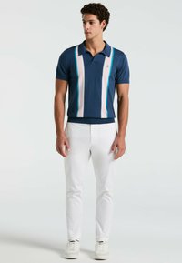 Marineblauwe poloshirt met verticale lichtroze en turkooise strepen, korte mouwen en een kraag, gecombineerd met witte slim-fit broeken en sneakers.