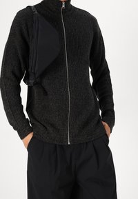 Knitted dark gray sweater with high collar and full zipper. Combined with loose black trousers and a black sling bag. Textured fabric.