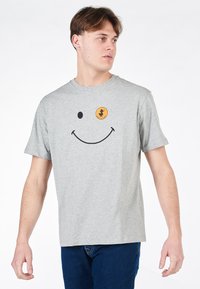 T-shirt in cotone grigio con grafica di un sorriso nero e un patch rotondo arancione con un design stampato. Maniche corte, vestibilità rilassata.