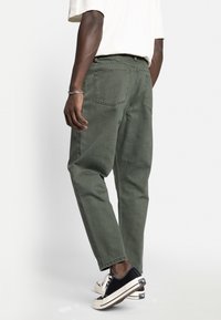Green cotton trousers with a straight fit, featuring back pockets and contrasting stitching, paired with black canvas sneakers.