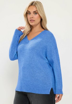 Femme blonde portant un pull bleu à manches longues col en V et un pantalon noir sur un fond uni, posant avec une main près de son visage.