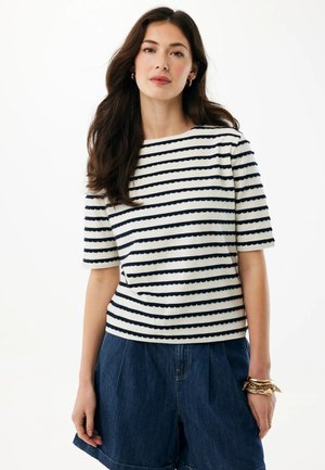 Vrouw met lang donker haar, gekleed in een wit kortemouwen shirt met getande marineblauwe strepen en losse donkerblauwe denimshorts, gecombineerd met gouden armbanden.