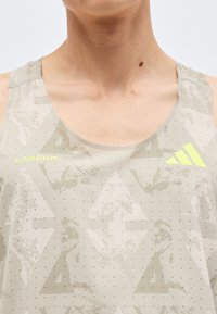 Κοντινό πλάνο ενός ατόμου που φοράει αθλητική μπλούζα αμάνικη σε ανοιχτό γκρι με αφηρημένα σχέδια, κίτρινο κείμενο "LÀBRUM" και κίτρινο λογότυπο Adidas.