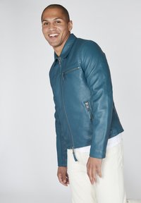 Giacca in pelle verde acqua con zip frontale, colletto e tasche con zip. Texture liscia e design aderente, indossata sopra una camicia bianca e pantaloni chiari.