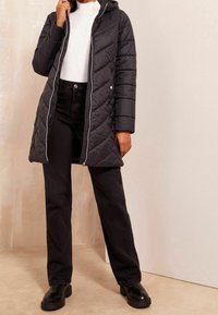 Osoba oblečená v černé prošívané dlouhé bundě s kapucí, bílém topu, černých kalhotách a černých kotníkových botách stojí uvnitř.