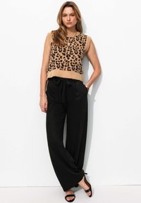 Pull sans manches marron clair avec un imprimé léopard noir, associé à un pantalon large noir et des sandales noires, avec une taille nouée.