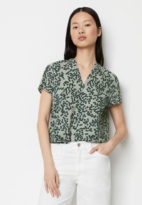 Kurzärmlige grüne Bluse mit V-Ausschnitt, die mit einem gepunkteten Muster in Dunkelgrün verziert ist. Weicher Stoff mit lockerer Passform, kombiniert mit weißen Hosen.