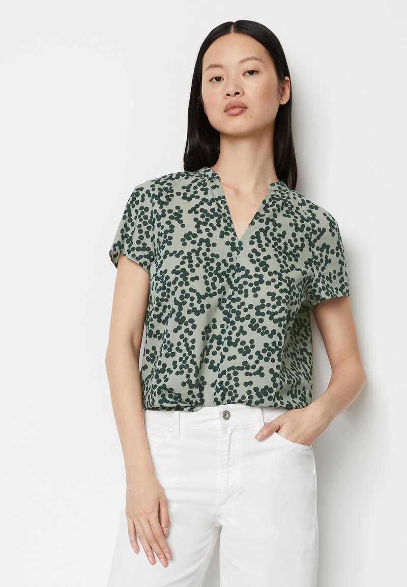 Kurzärmlige grüne Bluse mit V-Ausschnitt, die mit einem gepunkteten Muster in Dunkelgrün verziert ist. Weicher Stoff mit lockerer Passform, kombiniert mit weißen Hosen.