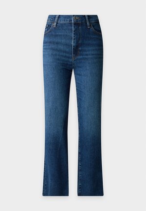 Femme aux longs cheveux noirs portant une chemise blanche sans manches boutonnée, un jean bleu et des talons hauts noirs, debout.