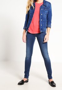 Chaqueta de denim en azul oscuro con botones plateados, combinada con una camisa roja y jeans ajustados azules. Mocasines negros completan el conjunto.