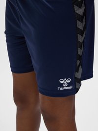 Marineblauwe sportshorts met zwart-witte chevronstreep aan de zijkant en wit hummel-logo nabij de zoom op de dij van een persoon.