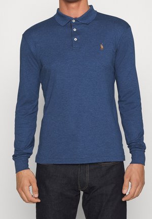 Homme portant un polo bleu à manches longues avec trois boutons et un jean foncé, debout devant un fond uni.