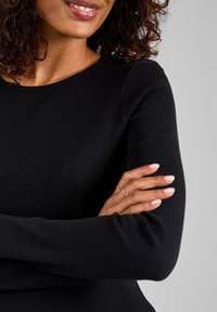 Žena s kudrnatými vlasy, která má na sobě černý top s dlouhými rukávy, lehce se usmívá, s překříženýma rukama a zlatými prsteny na prstech levé ruky.