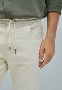 Pantalones beige claros con cinturilla elástica y cordón. Cuenta con bolsillos frontales y una textura suave. Los detalles incluyen herrajes plateados.