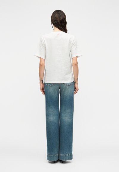 Λευκή κοντομάνικη βαμβακερή μπλούζα με κόκκινο λογότυπο στην πλάτη, σε συνδυασμό με ψηλόμεσα, φαρδιά τζιν παντελόνια σε μπλε χρώμα με ξεβαμμένο φινίρισμα.