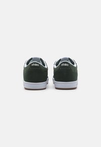Pantofi din suede verde închis cu accente albe, prezentând o vârf rotund, talpă albă și branding pe toc. Textură netedă și design casual.