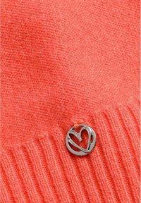 Heldere koraal gebreide stof met geribbelde rand, waaraan een kleine ronde zilveren charm met een hartvormig uitgesneden ontwerp is bevestigd.