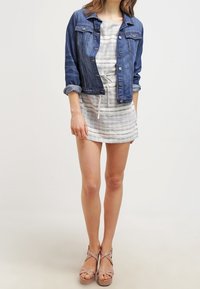 Tummansininen farkkutakki napittomilla kiinnityksillä ja käännetyillä hihoilla, kevyesti raidoitetun mekon päällä, jossa on kiristysnauha. Vaaleat korkeat sandaalit.