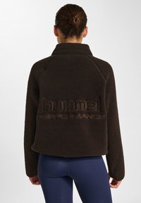 Brun fleecejacka med hög krage, har en djärv "hummel PERFORMANCE"-logotyp i svag kontrast, texturerad tyg och kort design.