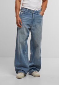 Világoskék denim anyagból készült, bőszárú farmernadrág, elhalványult felülettel. Öt zsebbel és gombos záródással rendelkezik. Fehér sportcipővel viselve.