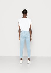 Top branco sem mangas e jeans claros de corte slim. Os ténis são brancos. O fundo é simples e neutro.