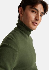 Suéter verde de cuello alto, hecho de un tejido de punto suave y texturizado. Presenta un cuello alto y una silueta ajustada, mostrado desde un ligero ángulo lateral.