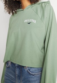 Zelená zkrácená mikina s kapucí, se bílým textem a logem "HOLLISTER". Vyrobena z měkkého materiálu, má dlouhé raglánové rukávy.