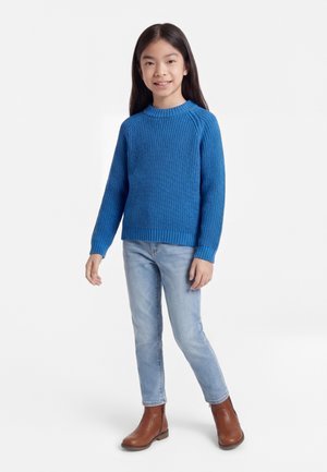 Jeune fille debout, portant un pull en maille bleu, un jean bleu clair et des bottines marron, sur un fond blanc uni.