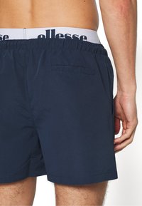 Marineblå svømmebukser med elastisk talje og et mærketikette. En bagside lomme er synlig på højre side. Glat, letvægtsstof.