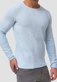 Pull-over bleu clair à manches longues avec un tissu en tricot texturé. Encolure arrondie et manches ajustées. Pantalons gris avec une main dans la poche.