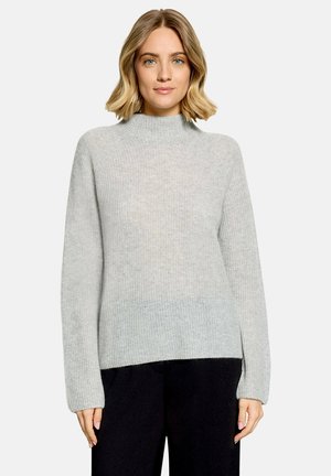 Femme aux cheveux blonds mi-longs portant un pull en maille gris clair à col montant et un pantalon noir, debout devant un fond uni.
